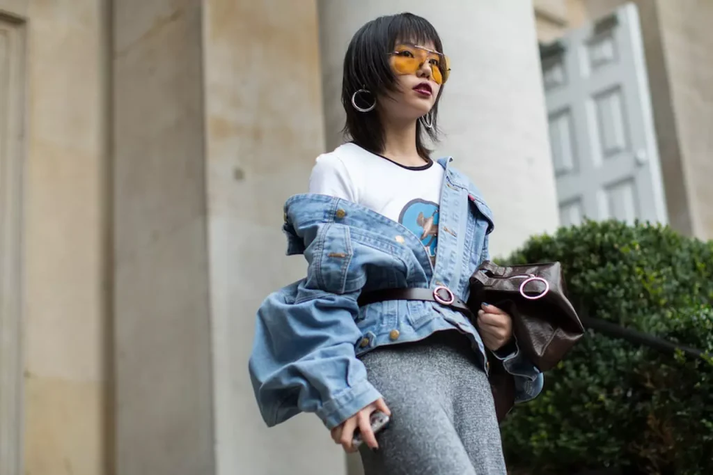 modelo usando jaqueta jeans oversized com cinto em couro marcando a cintura, saia em malha e óculos com lentes amarelas