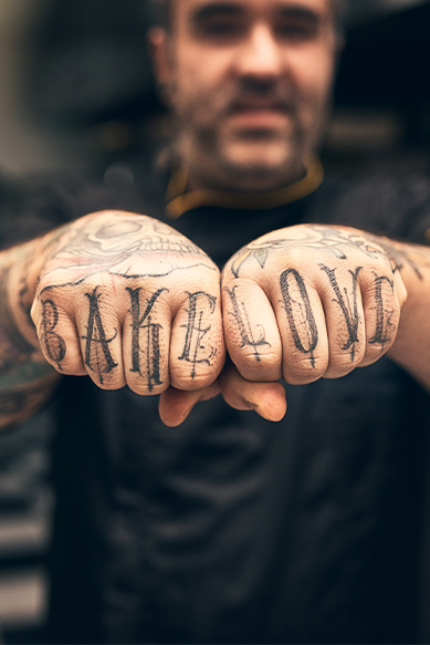 Homem com tatuagens nas mãos mostrando a frase 'Bake Love'